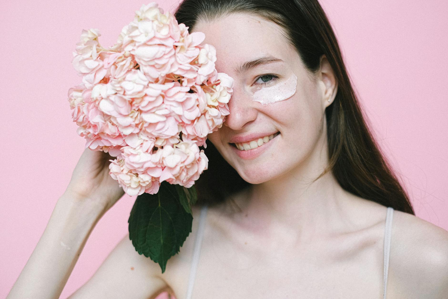smiling woman covering eye with pink flower while standing against pink background